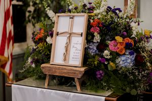 A wooden frame with a cross stands on an easel surrounded by vibrant floral arrangements with red, purple and blue blooms.
