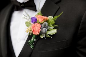 Close-up of wedding boutonnieres with coral roses, purple flowers, and green foliage pinned to black lapels.
