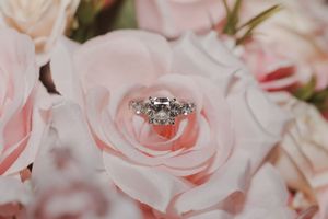 A diamond engagement ring nestles in the center of a soft pink rose bloom among pink and white floral arrangements.