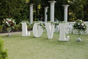 Large white LOVE letters decorated with flowers and greenery sit on grass with elegant columns in a garden wedding setup.