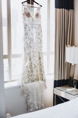 An elegant white lace wedding gown with floral embroidery hangs against a window with two-tone curtains.