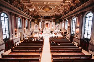 View down the center aisle of an ornate Catholic church with wooden pews and baroque architectural details.