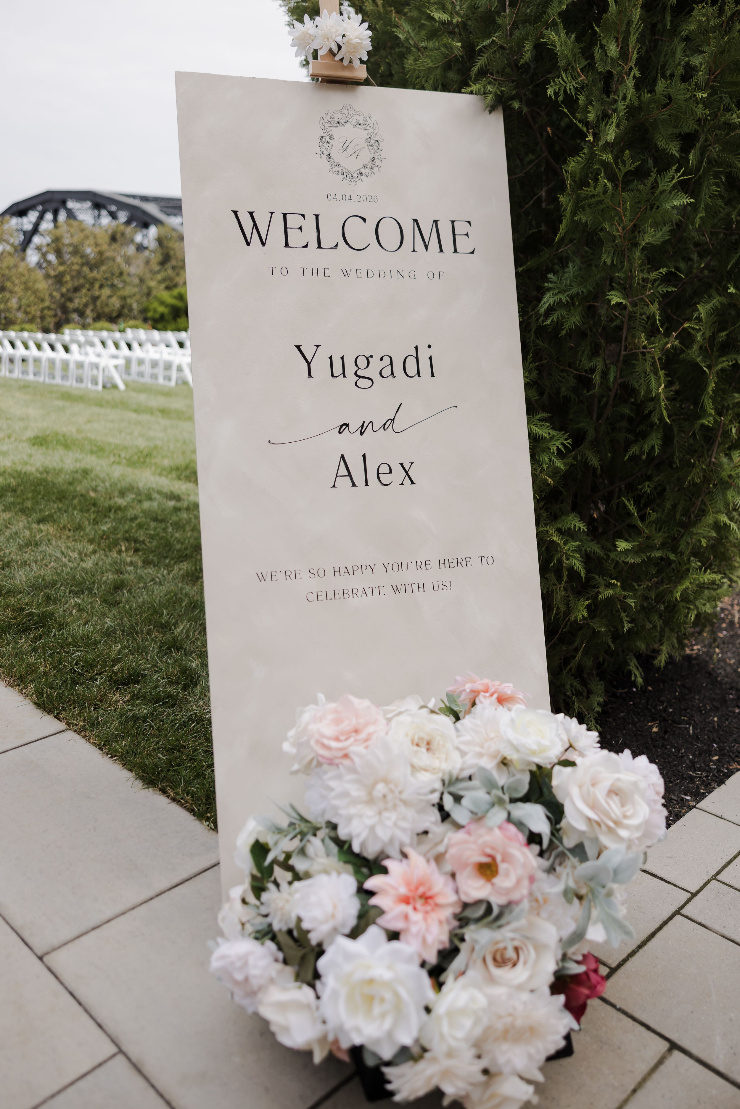Wedding welcome sign for Yugadi and Alex adorned with white and pink floral arrangement on a paved outdoor venue.