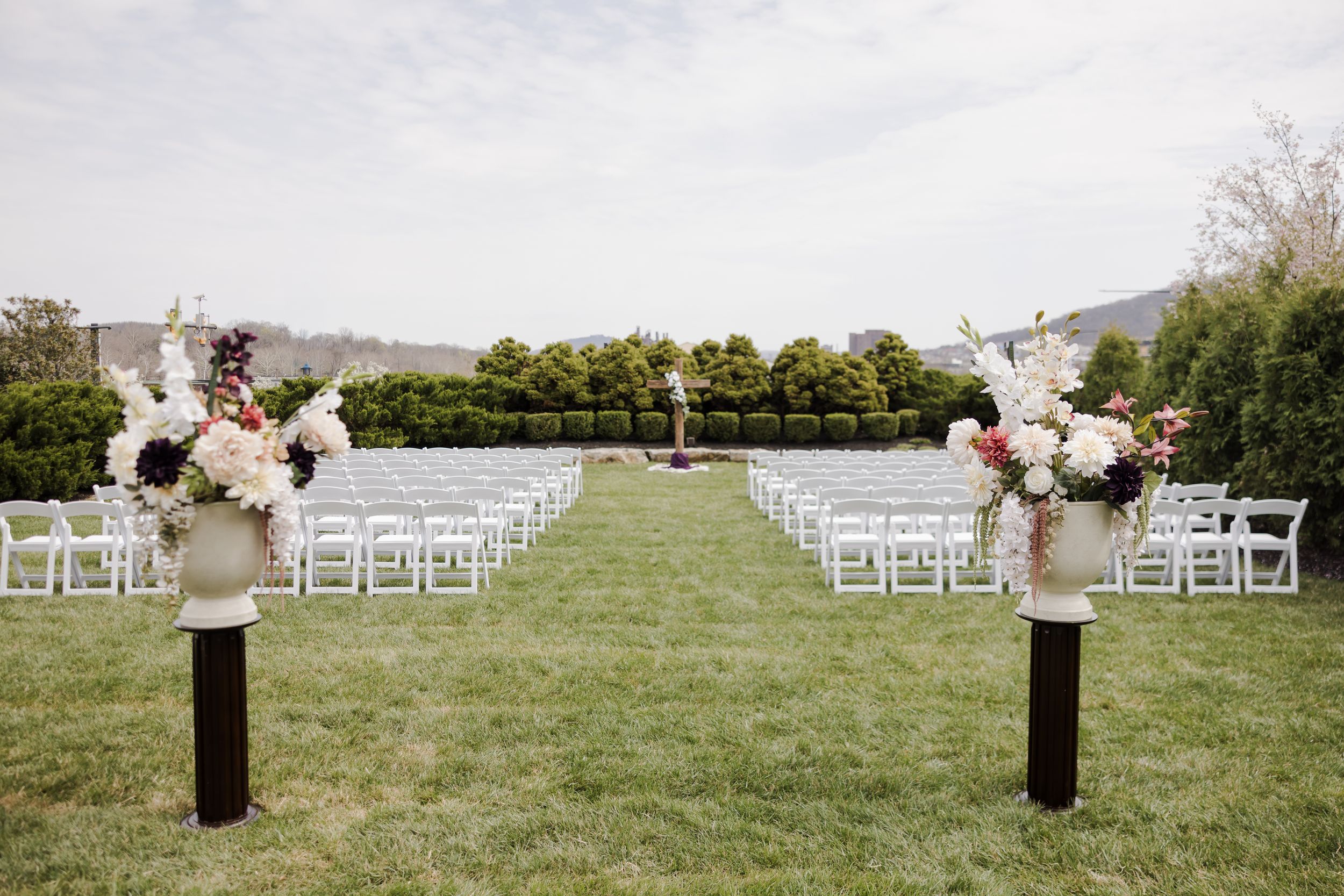 Elegant outdoor wedding ceremony setup with white chairs, floral arrangements on pedestals, and lush green hedges backdrop.