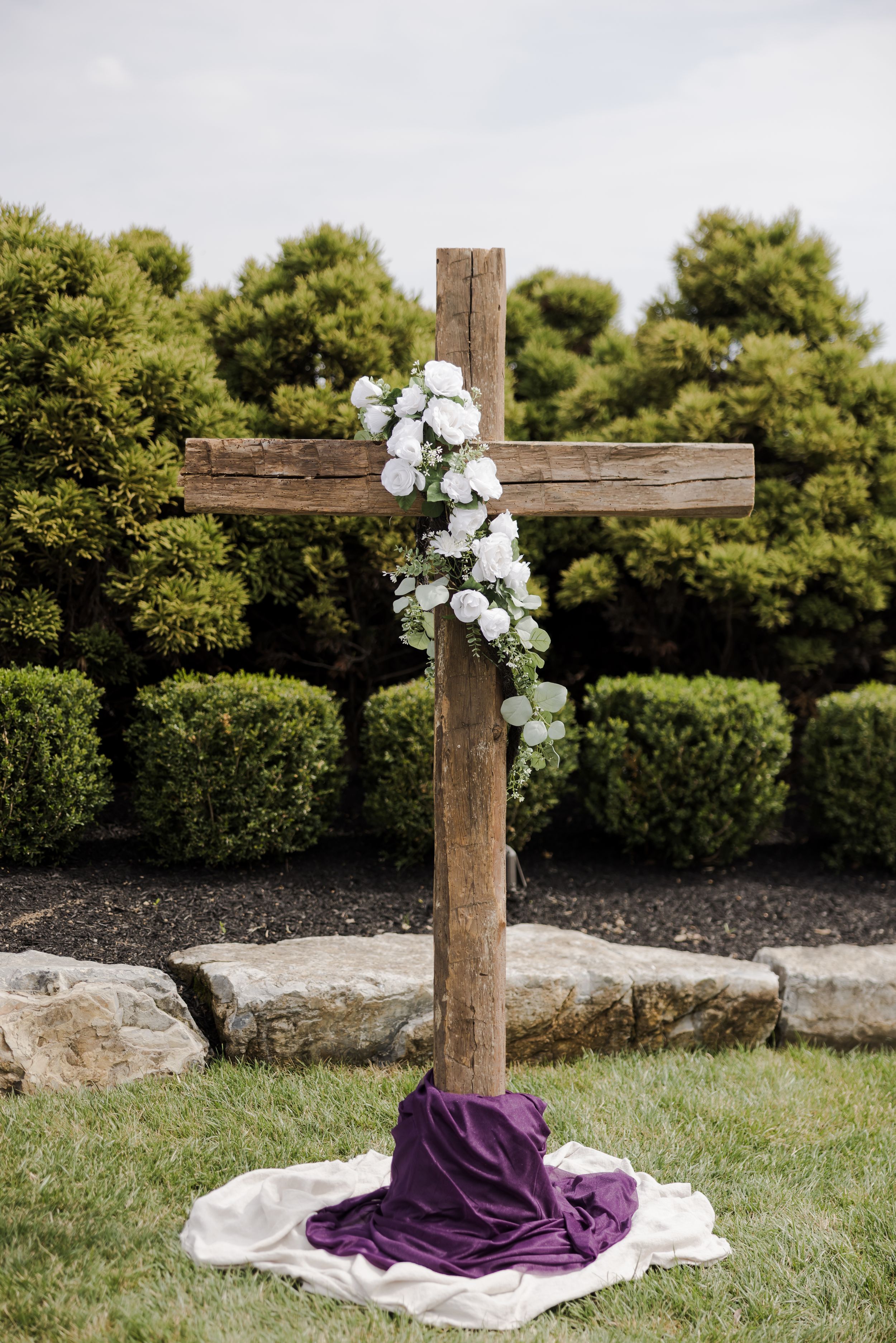 Rustic wooden cross decorated with white roses and purple drape on white cloth, set in a garden with trimmed hedges.