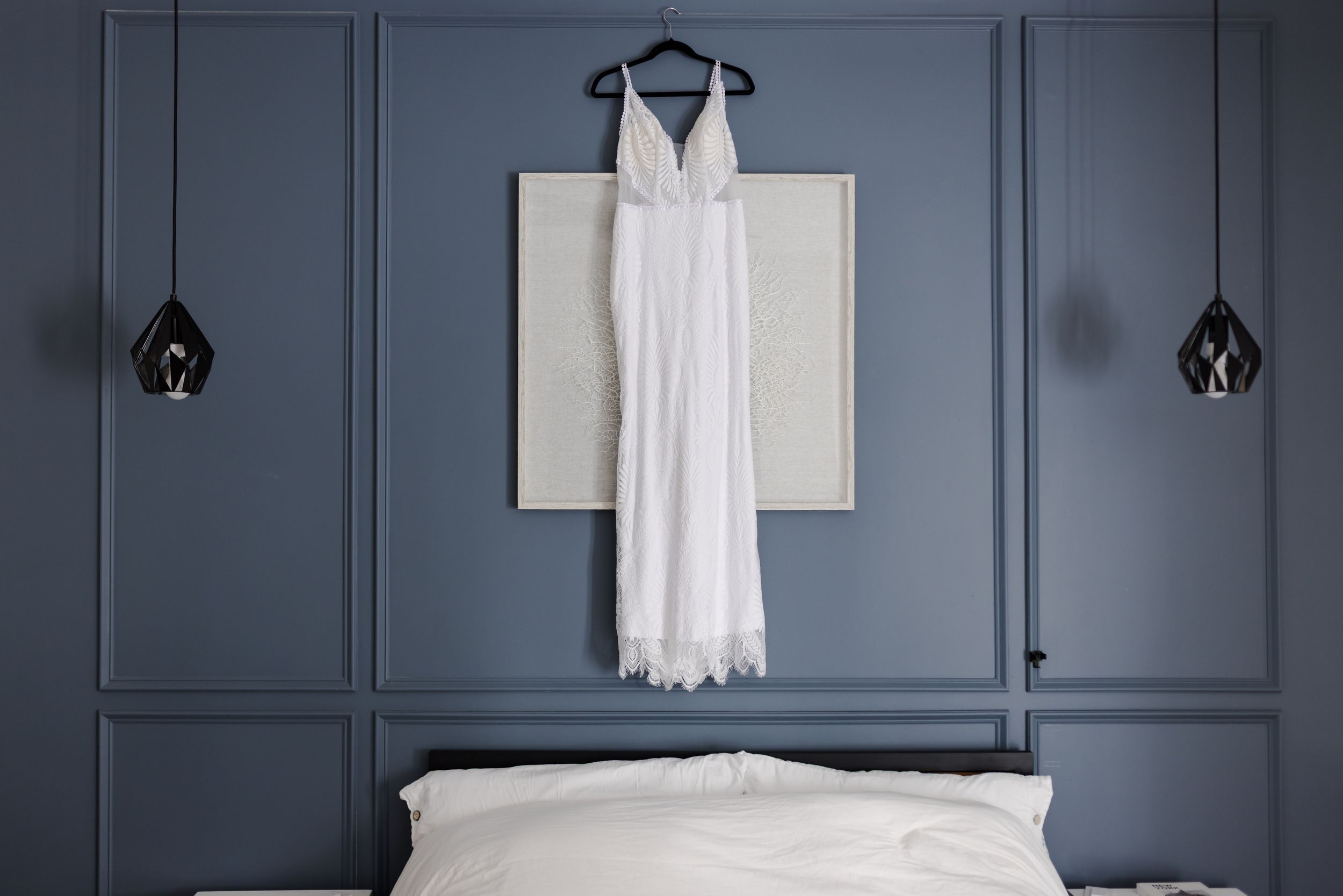 A modern bedroom features a white bed against dark blue paneled walls with white decor and pendant lights.