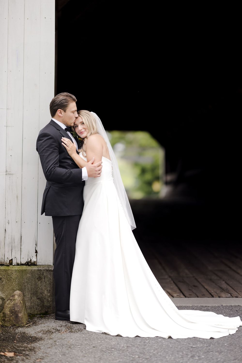 A romantic wedding couple embracing in front of a rustic white wooden barn backdrop.