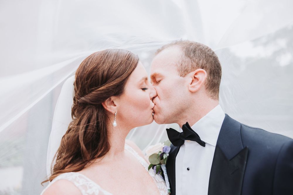 A romantic kiss between newlyweds outdoors with the bride wearing a veil and groom in classic black tuxedo.