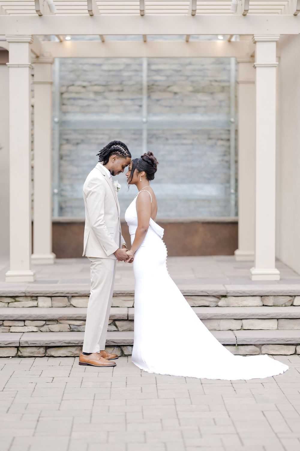 A couple in white wedding attire share an intimate moment under columns on a stone patio during their special day.