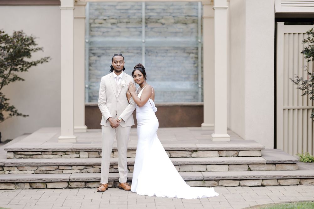 A wedding couple relaxes on stone steps in front of a stately white columned entrance with a lakefront view.