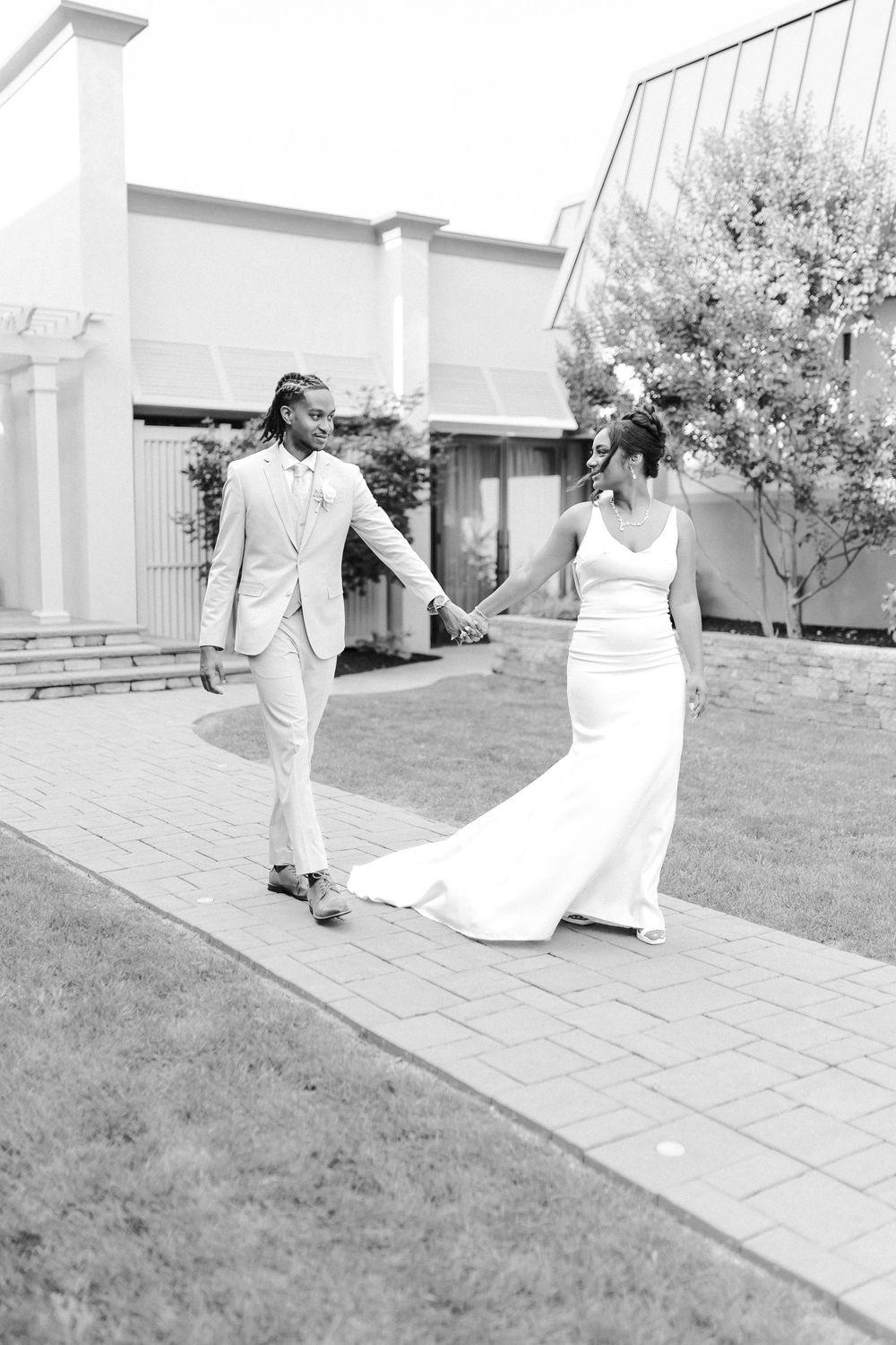 A couple in wedding attire walks hand in hand along a patio pathway in a black and white photo.