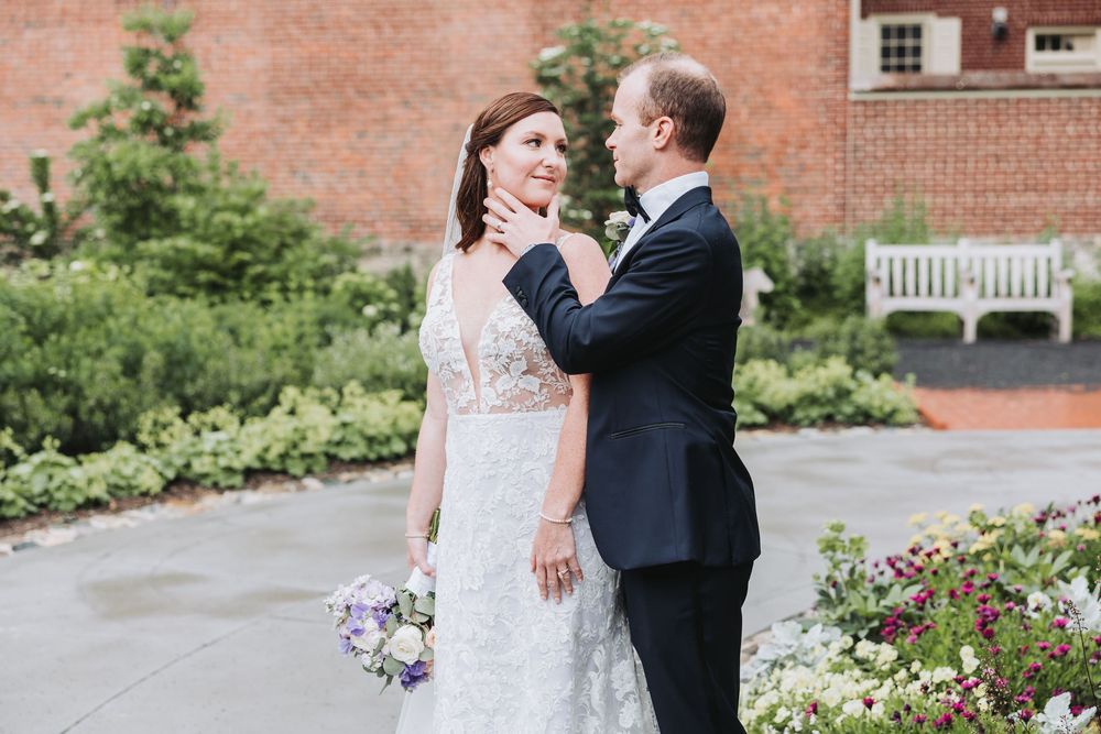A couple shares a romantic moment in their wedding attire near a garden path with brick walls and flowers.