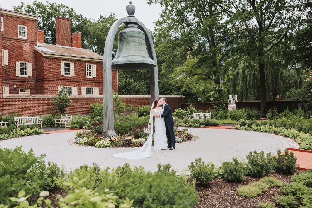 A couple stands under an ornate garden bell structure surrounded by manicured landscaping and brick buildings.