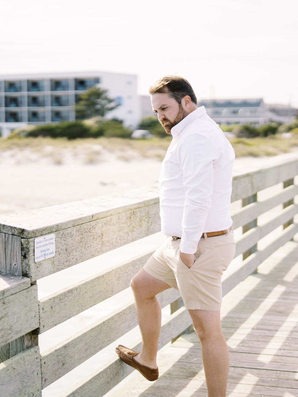 A person in summer attire stands on a wooden boardwalk near the beach.