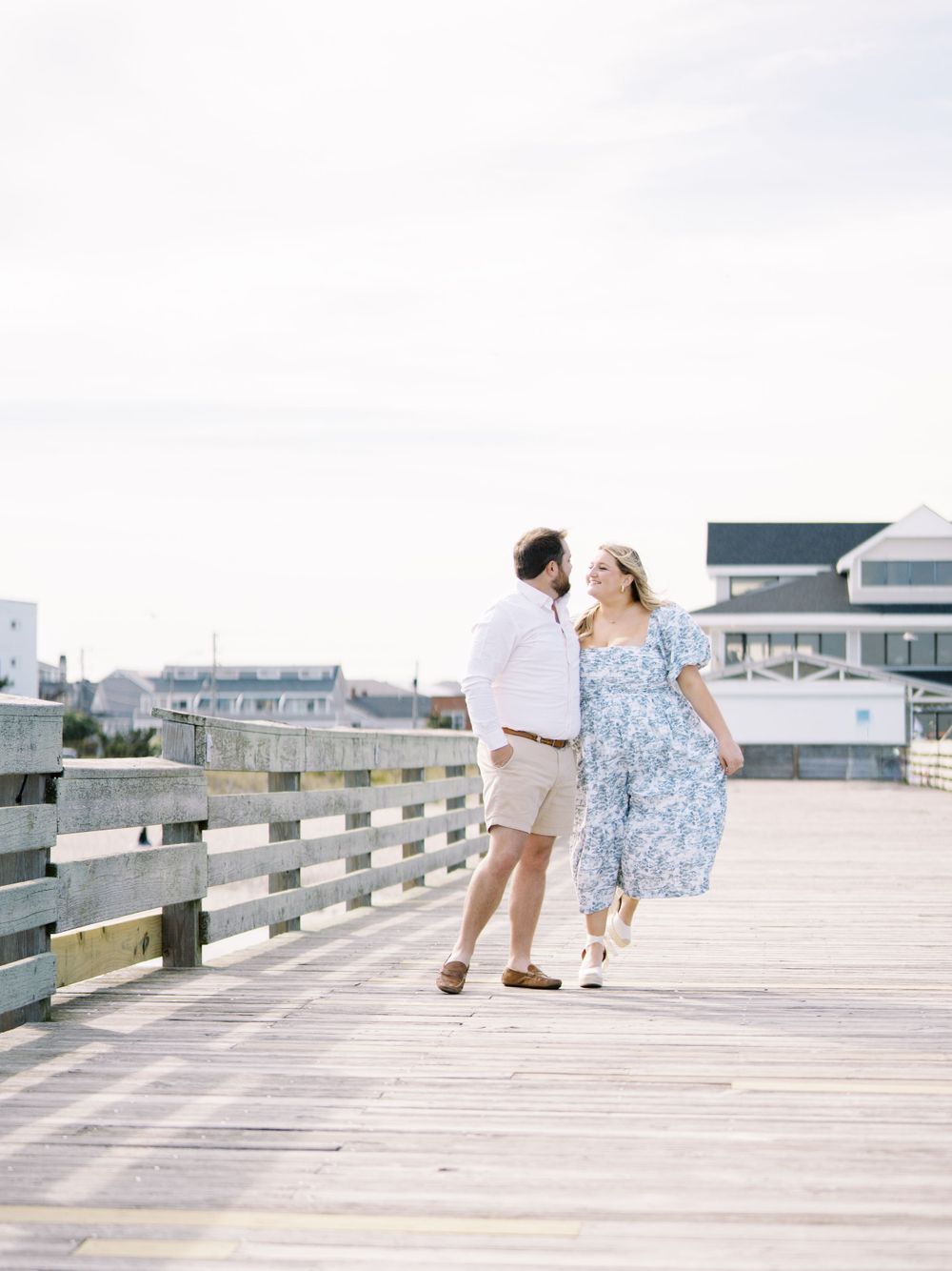 A couple in summer attire sharing a romantic moment on a wooden boardwalk by the ocean.