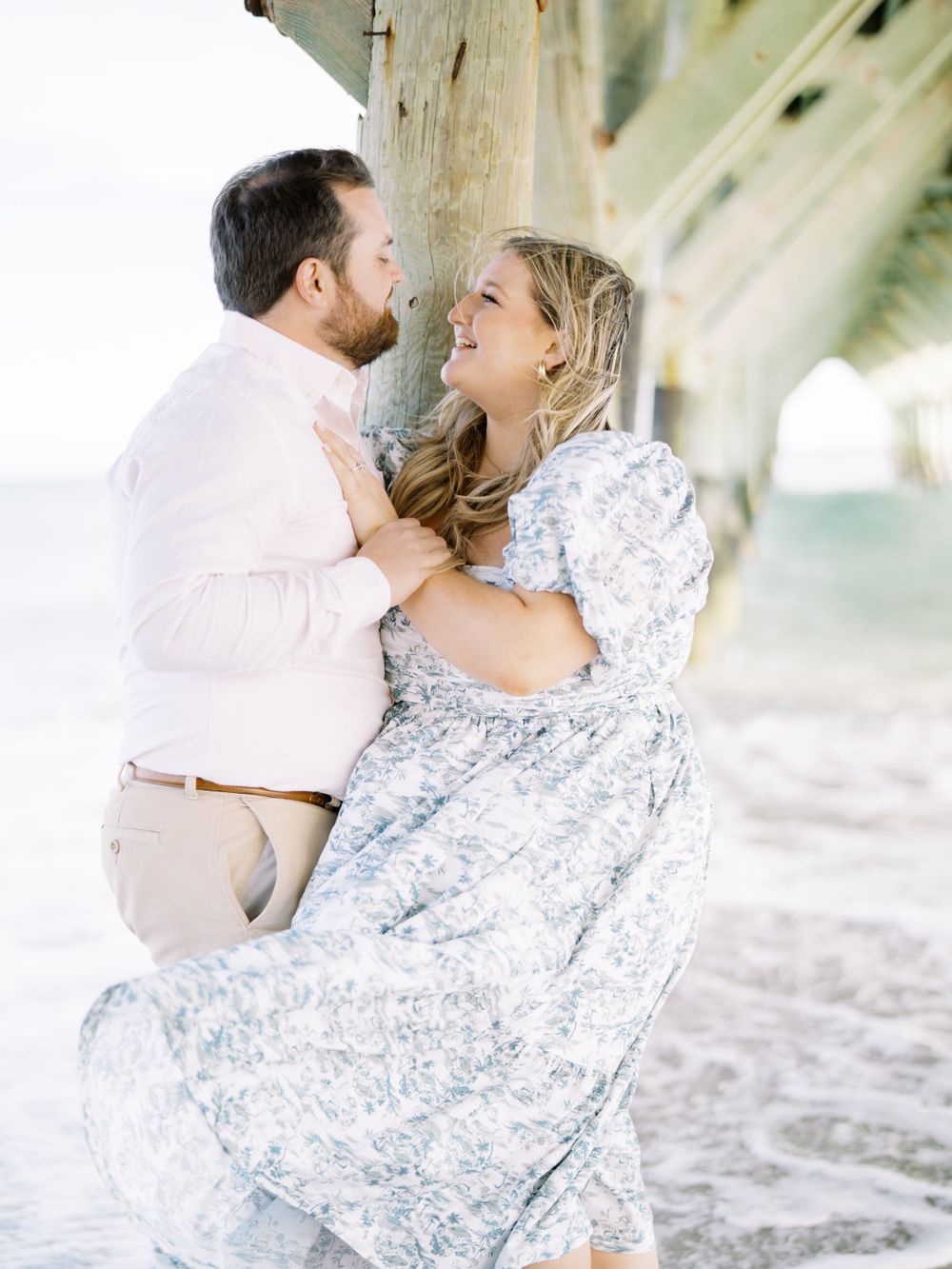 A romantic embrace under a beach pier with waves in the background.