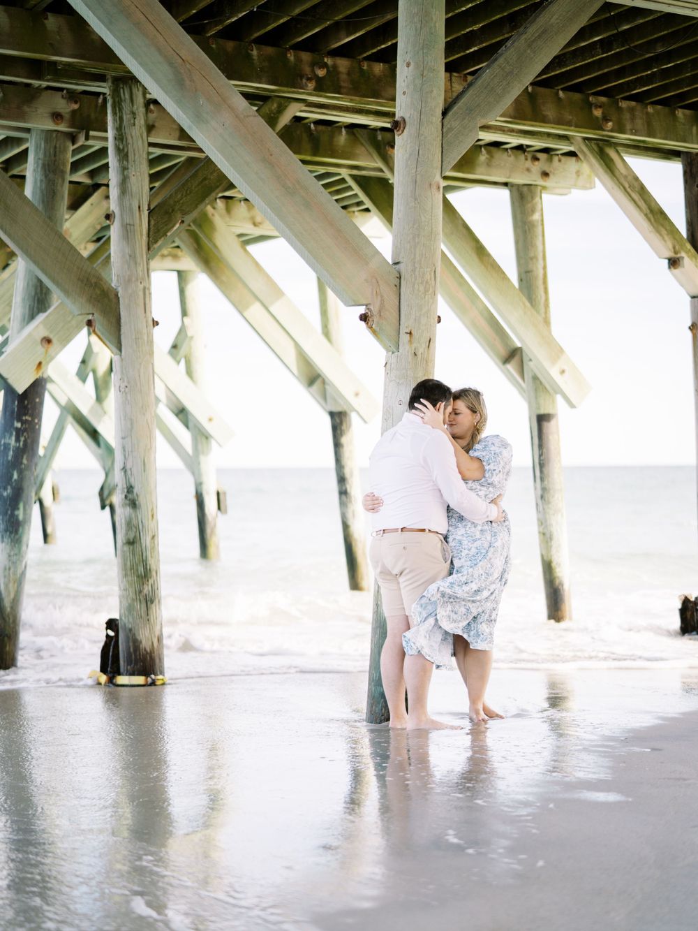 A couple sharing an intimate moment beneath a weathered wooden pier by the ocean.