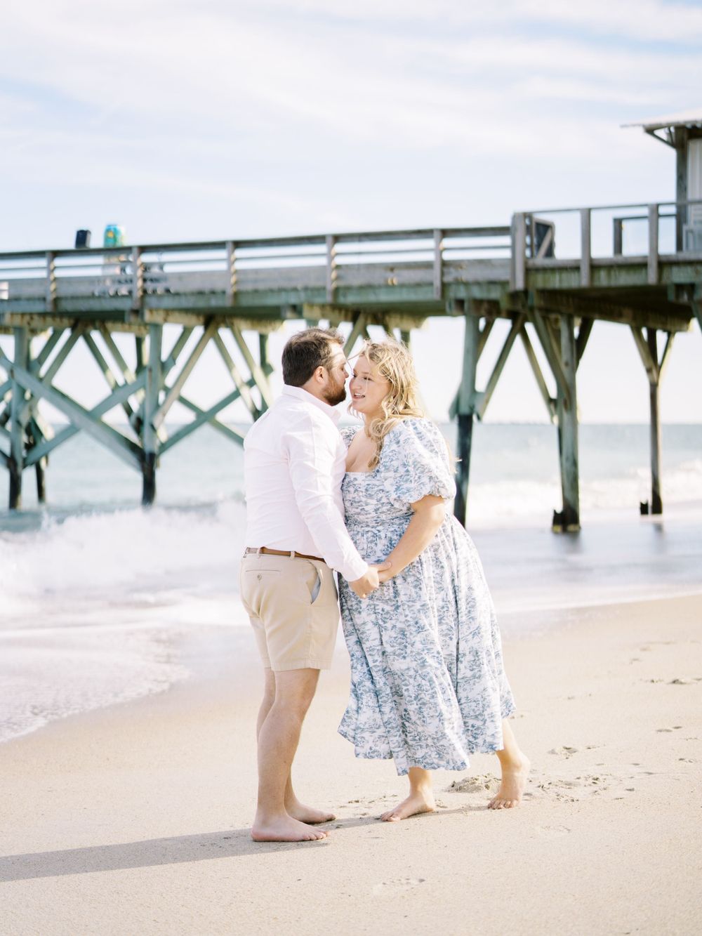 A couple embracing by a wooden pier at the beach during a romantic photoshoot.
