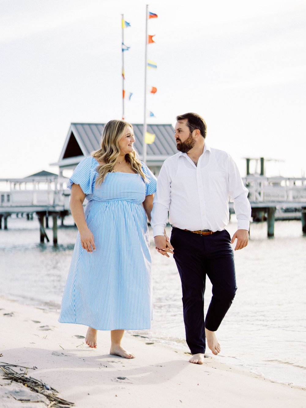 A couple walking hand in hand on a beach by a pier with colorful flags flying overhead.