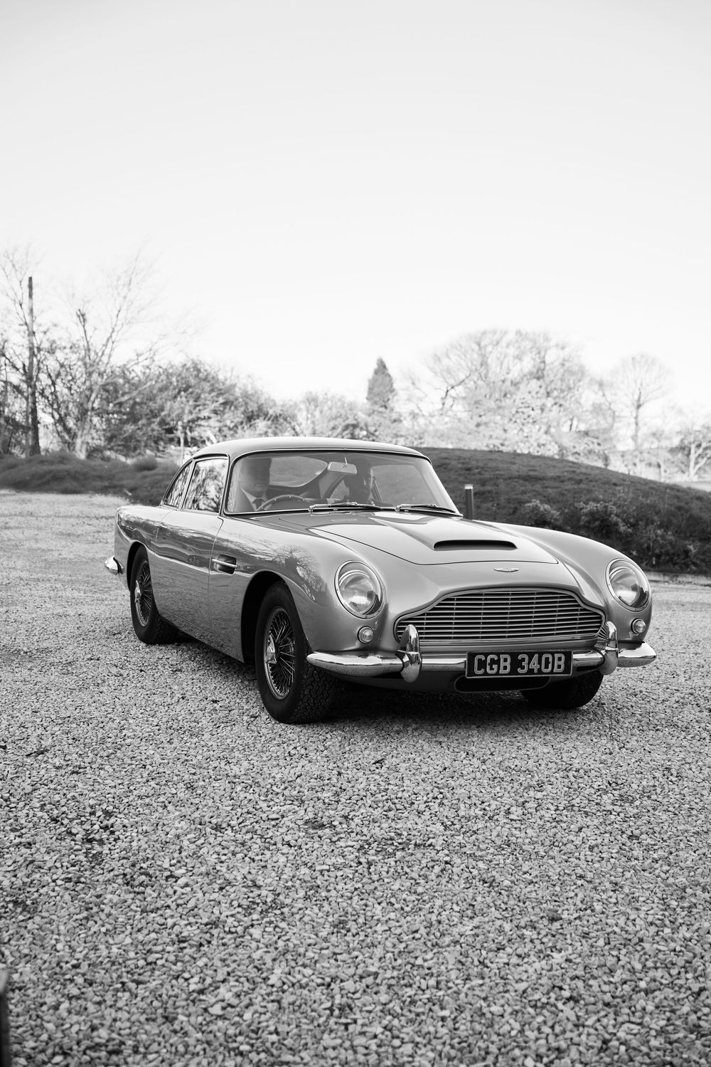 Classic Aston Martin DB5 sports car photographed on gravel in black and white.
