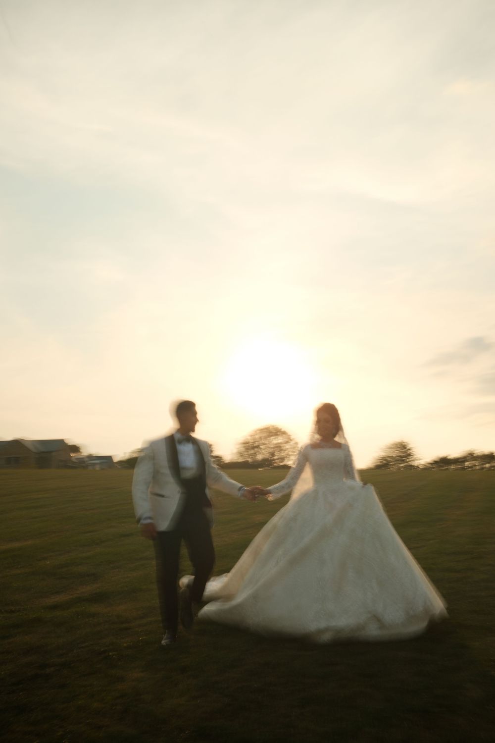 A couple in wedding attire walks across a grassy field at sunset creating a romantic silhouette.