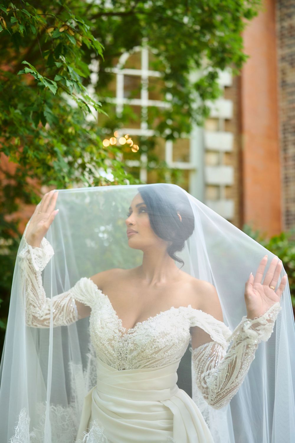 Ethereal bridal portrait through delicate white veil against green foliage background.