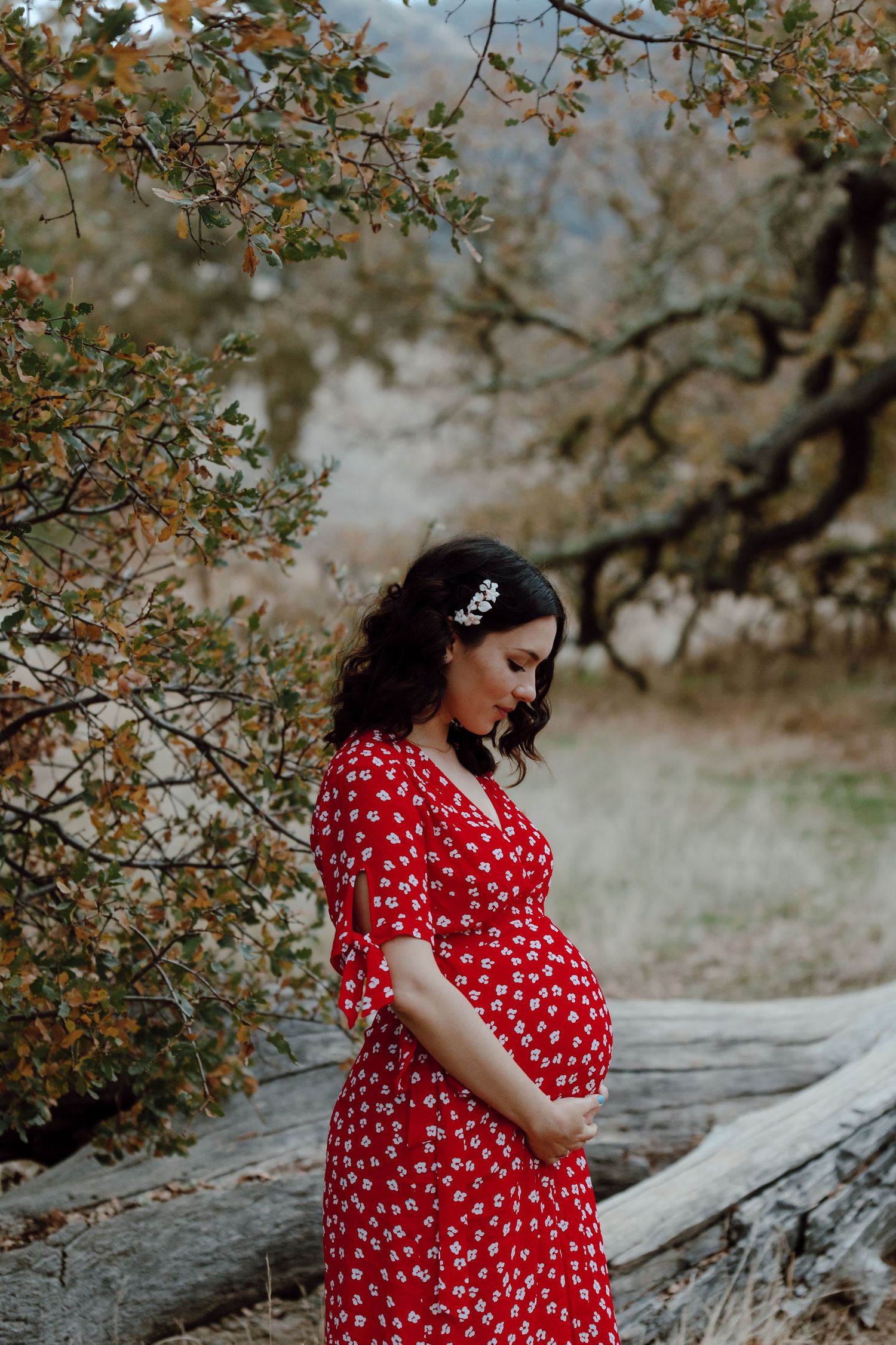A pregnant woman in a red floral dress poses outdoors under blooming tree branches.