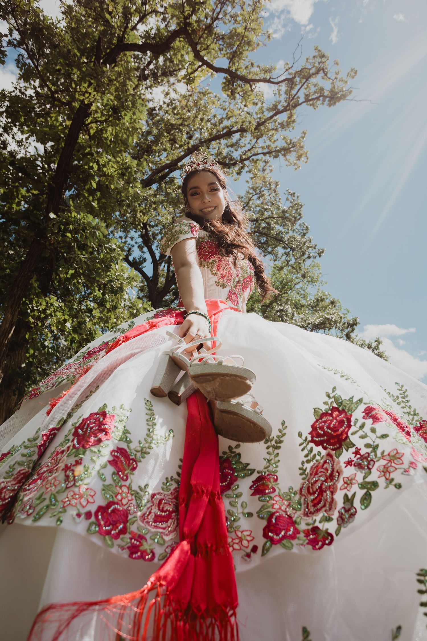 An ornate white and red floral dress photographed from below against a sunny blue sky and green trees.