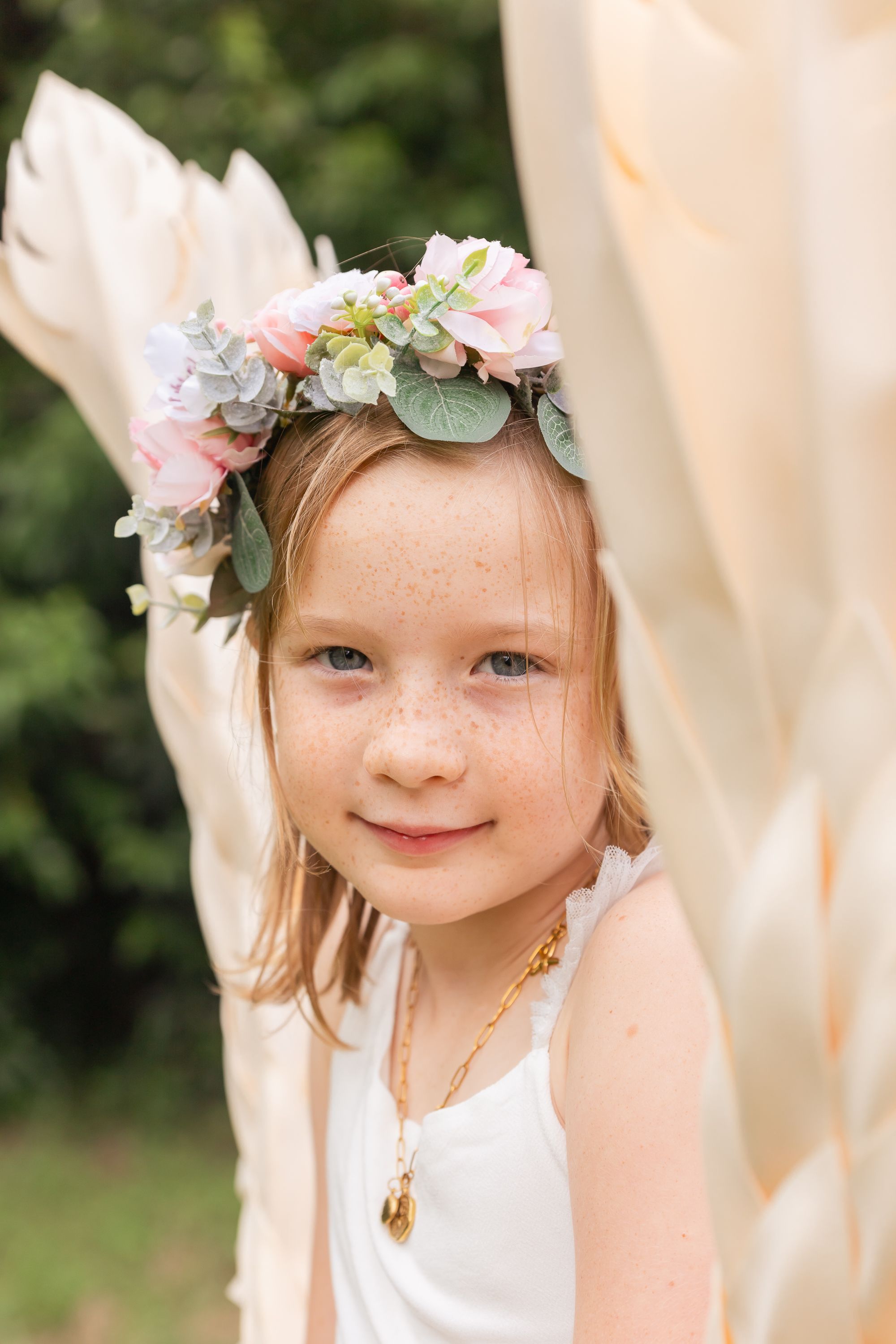 Young child wearing a white dress and floral crown smiles sweetly in natural outdoor lighting.
