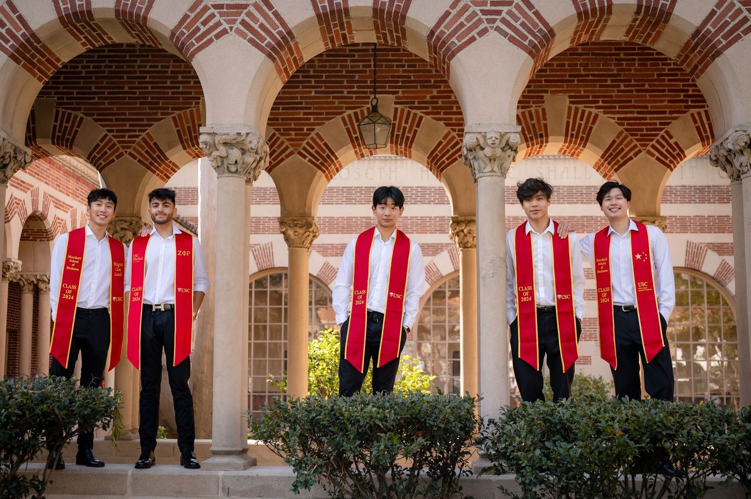 Five graduates in black suits and red stoles pose beneath ornate stone archways at a university courtyard.