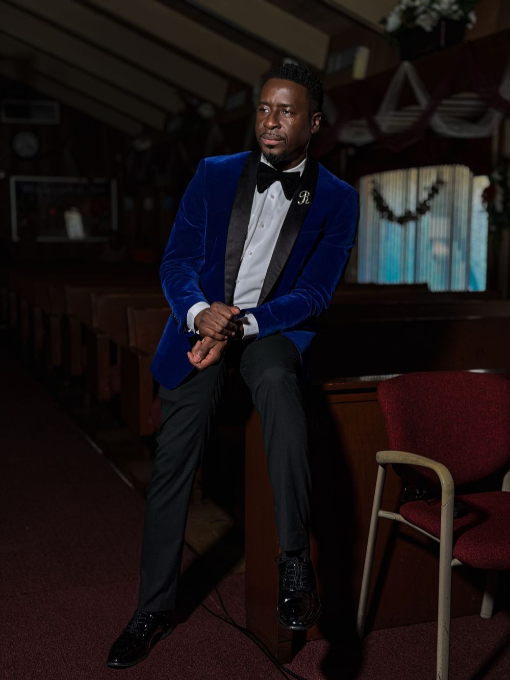 A well-dressed person in a royal blue velvet tuxedo jacket and black bowtie poses on a chair in a dimly lit room.