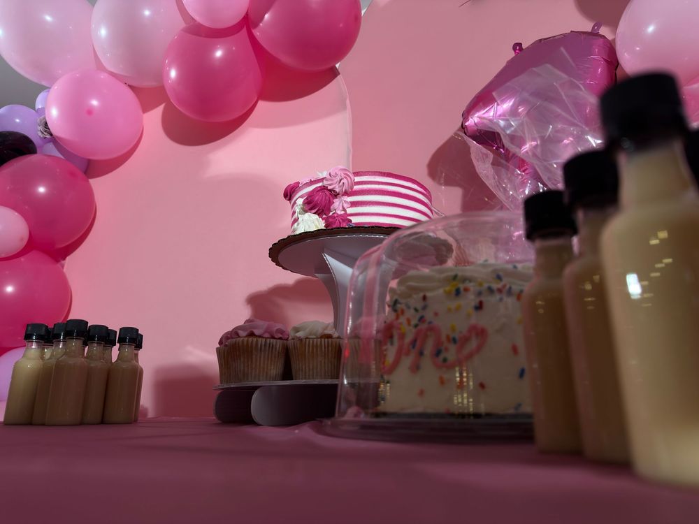 Pink balloon garland and dessert table setup with cake stand and bottles at a party celebration.