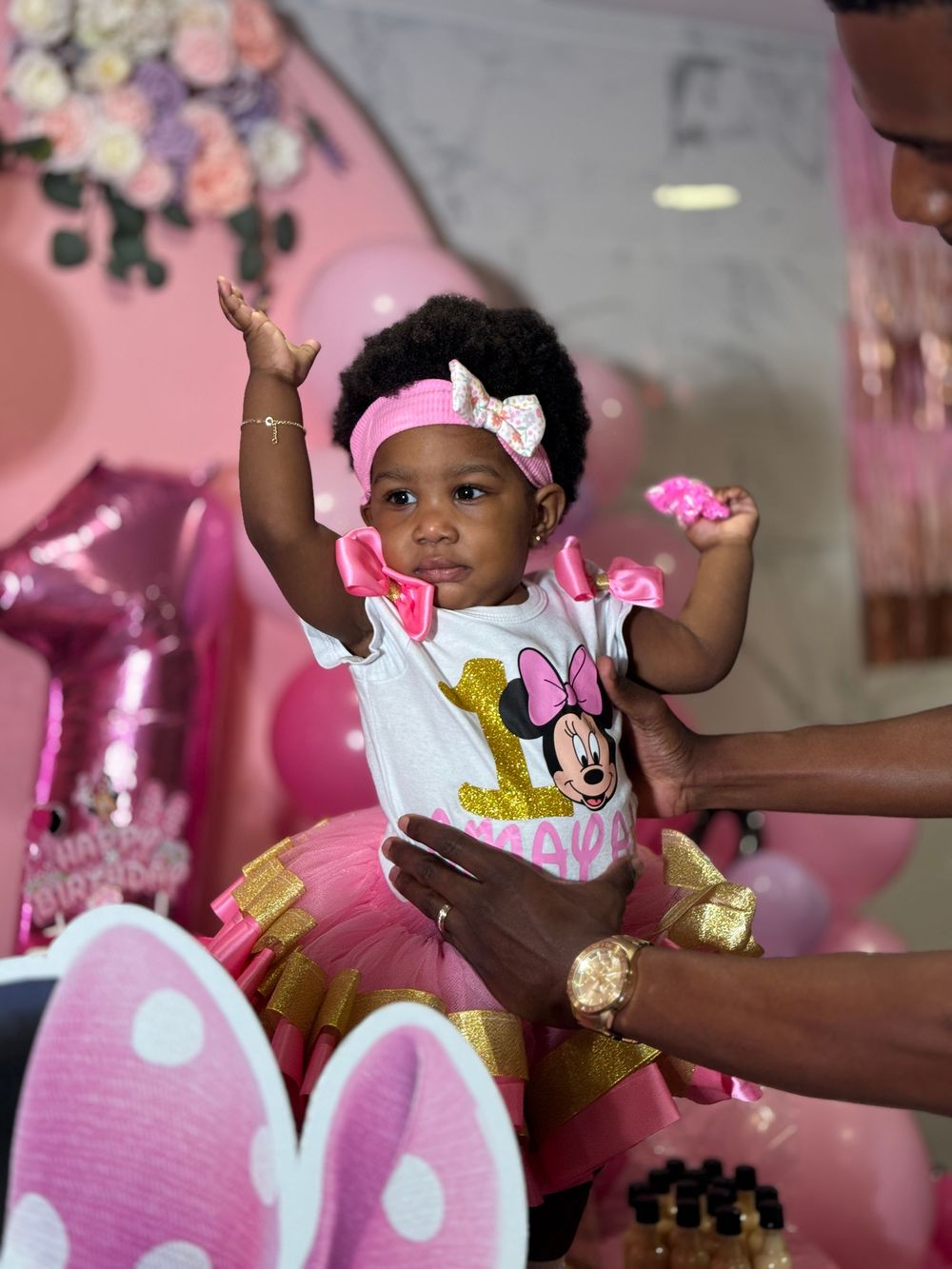 Small child in Minnie Mouse themed party outfit with pink and gold tulle skirt raises arms celebrating at festive pink event.