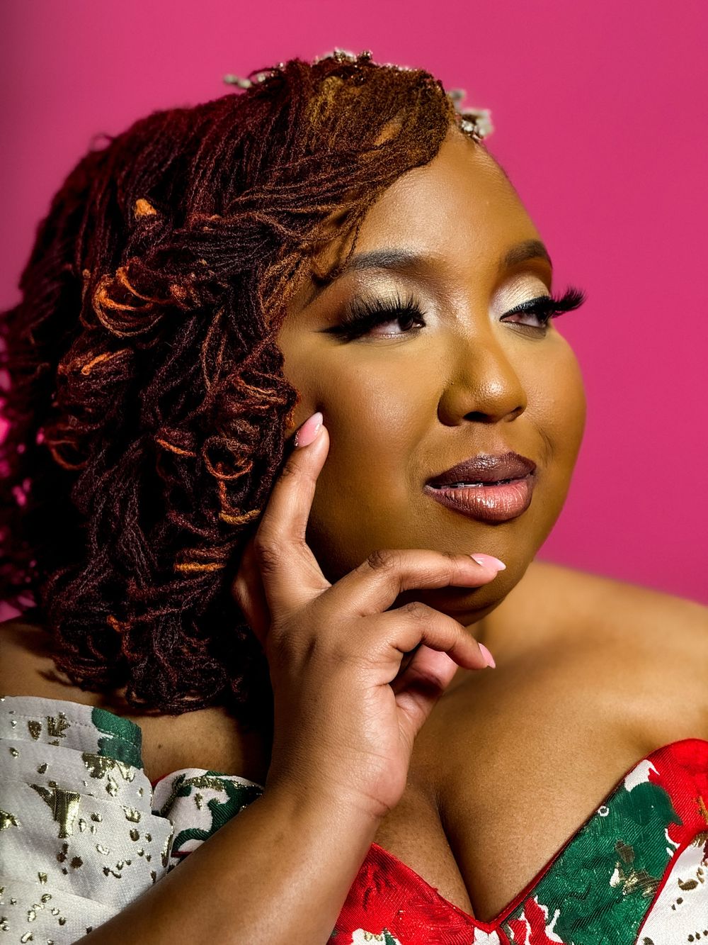 Beautiful portrait against pink background featuring red dreadlocks, glamorous makeup and red and green floral attire.