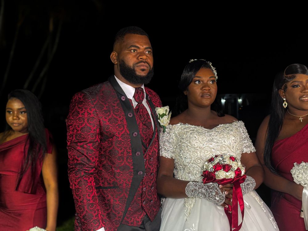 Wedding party stands together at night with the groom in a burgundy suit and bride in white gown holding red bouquet.