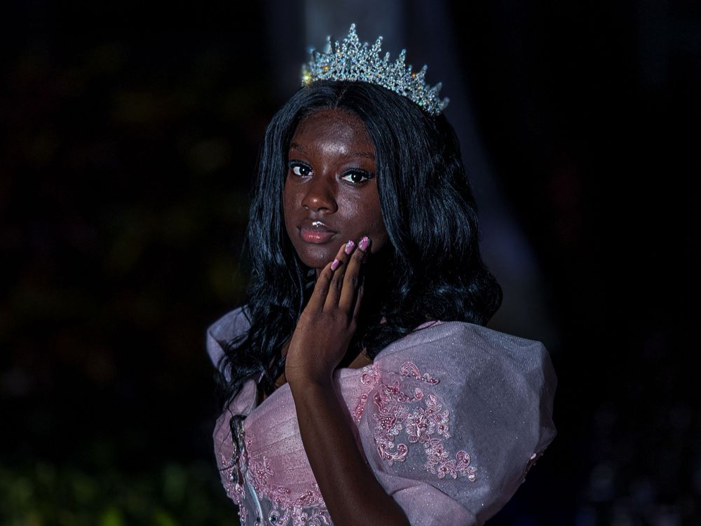 A person wearing a pink gown and sparkling tiara poses elegantly against a dark background.