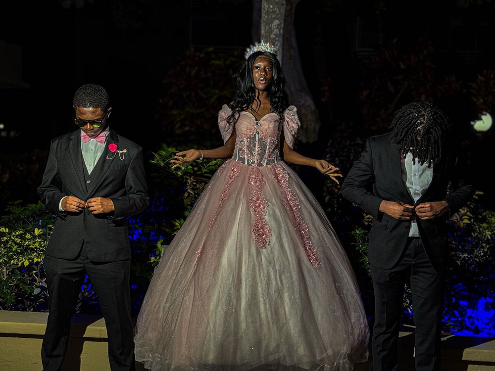 A person in a pink quinceañera ball gown stands between two attendants in dark suits during an evening celebration.