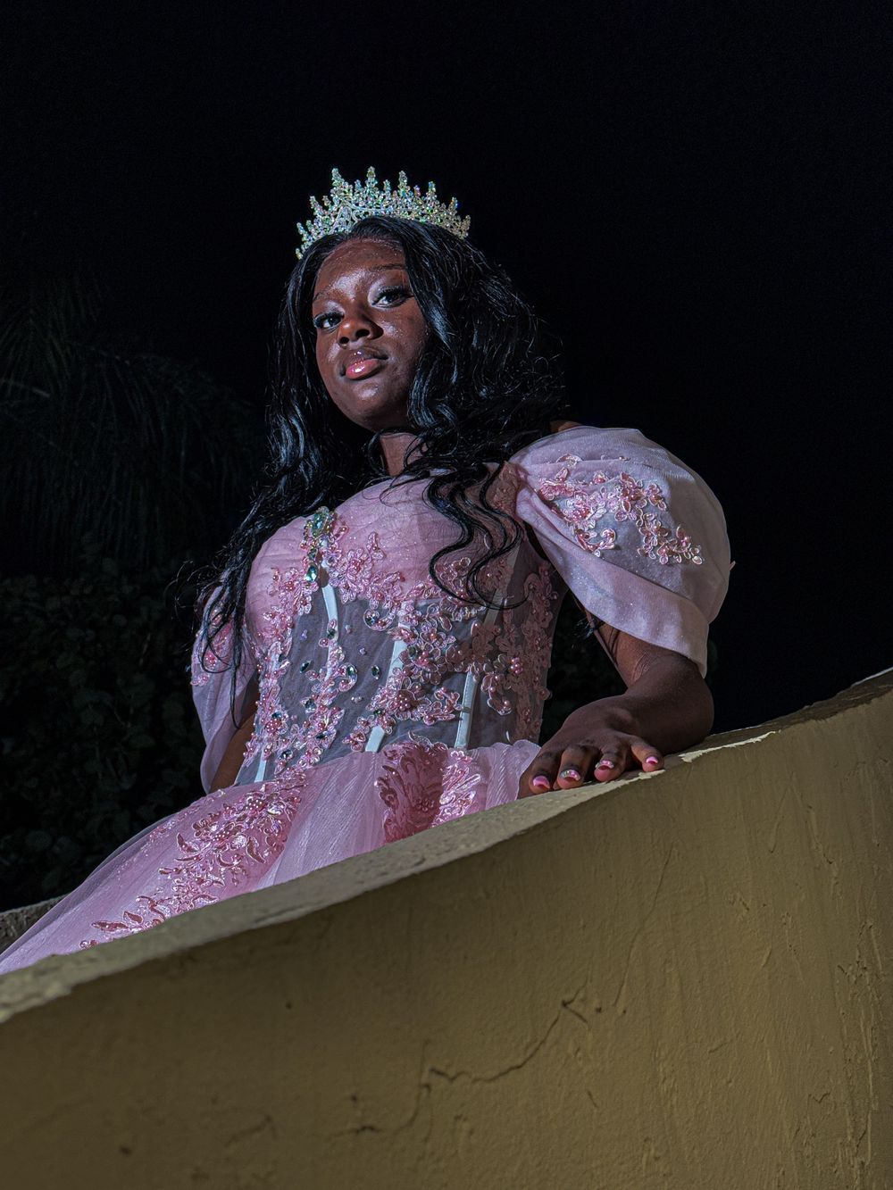A person wearing a sparkling pink ball gown and crystal crown posed at night against a dark background.