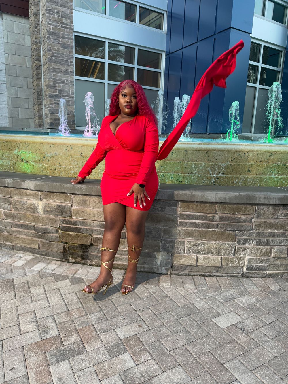 A vibrant red dress and matching hair color stands out against stone architecture and water fountains in an outdoor setting.