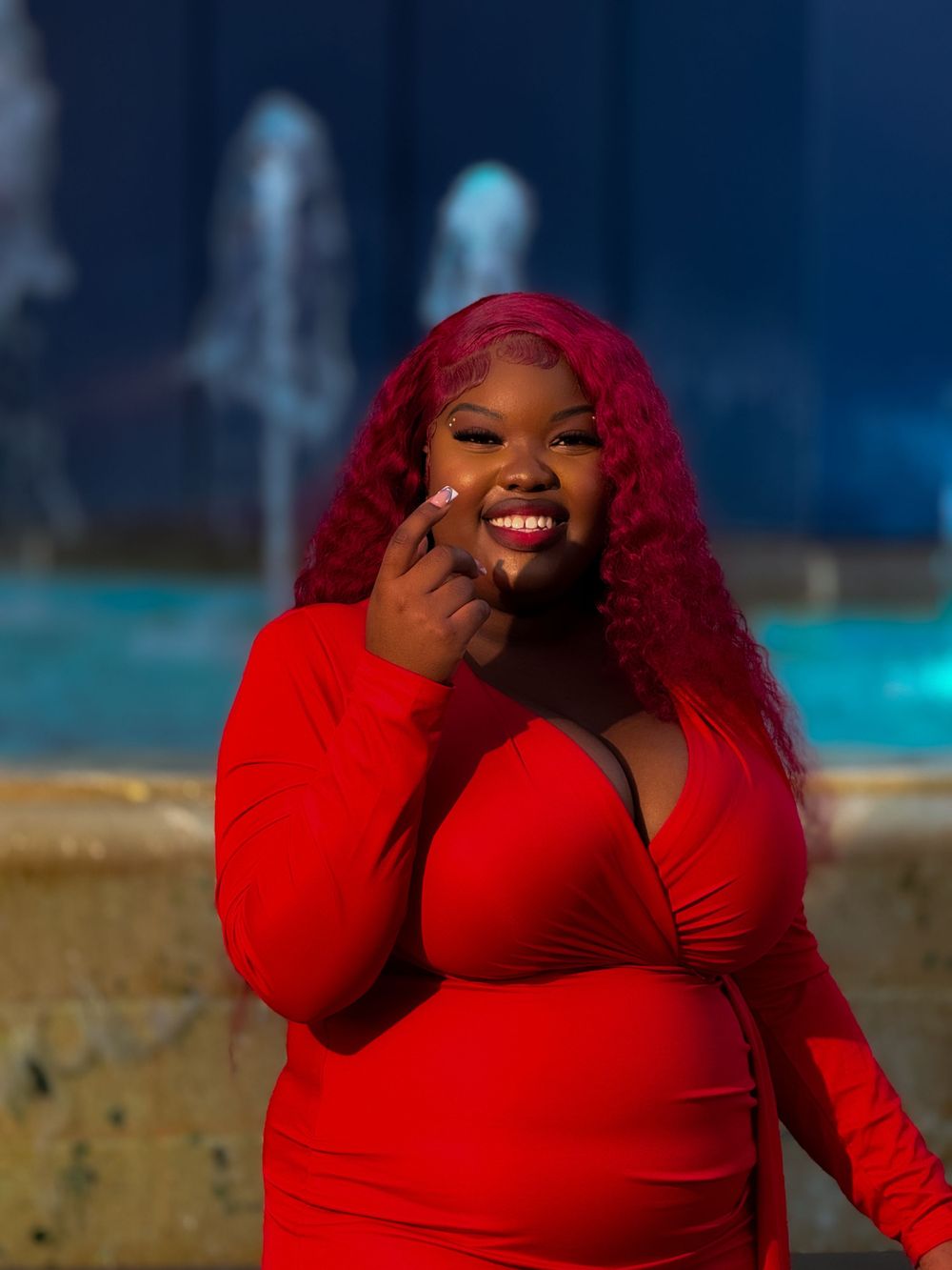 Person with vibrant red curly hair wearing a red long-sleeve dress poses outdoors by a pool at night.