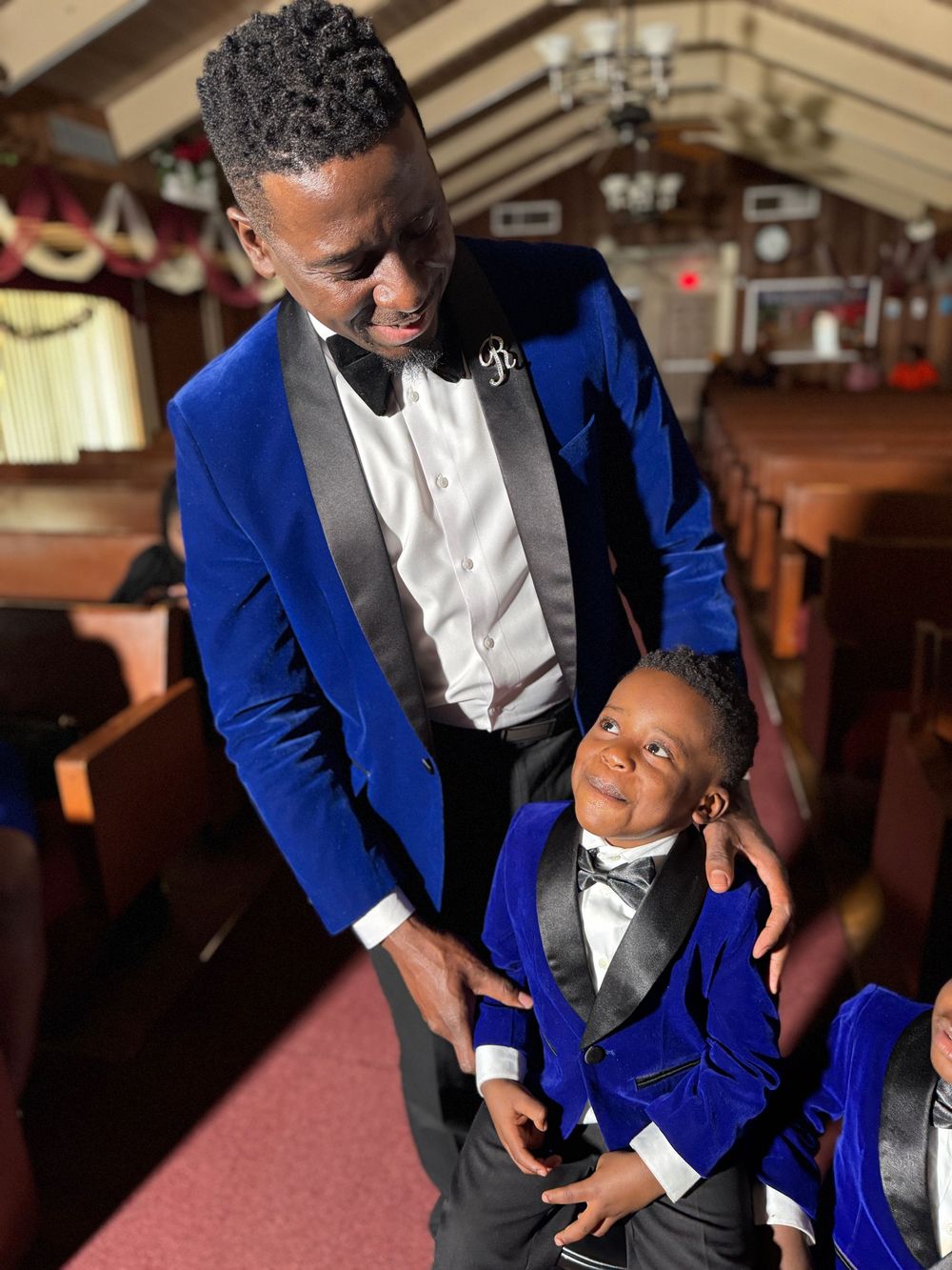 A pair in matching royal blue tuxedo jackets and black bow ties share a tender moment in an indoor venue.