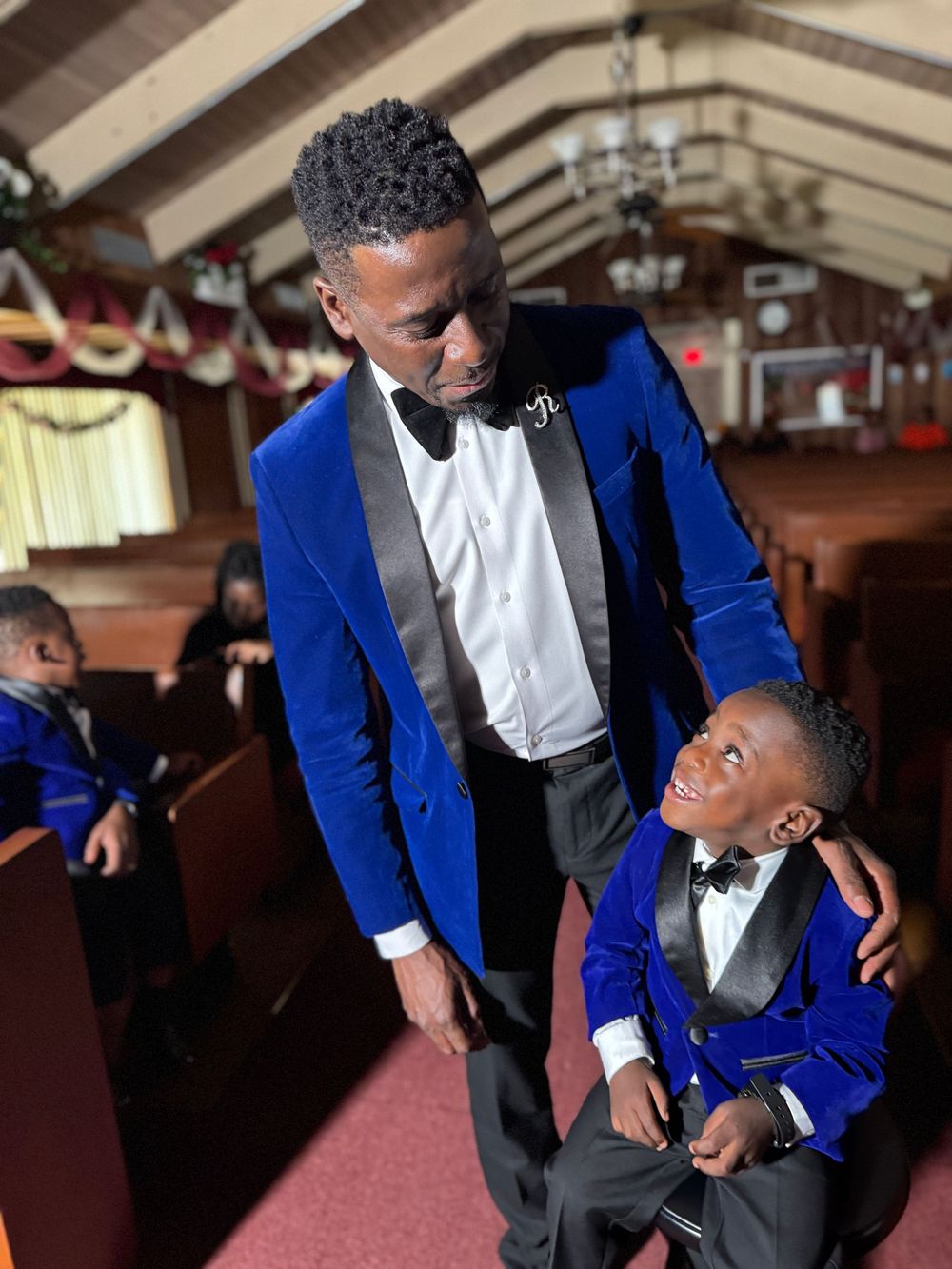 A touching moment showing matching royal blue velvet tuxedos during a formal event in an ornate venue.