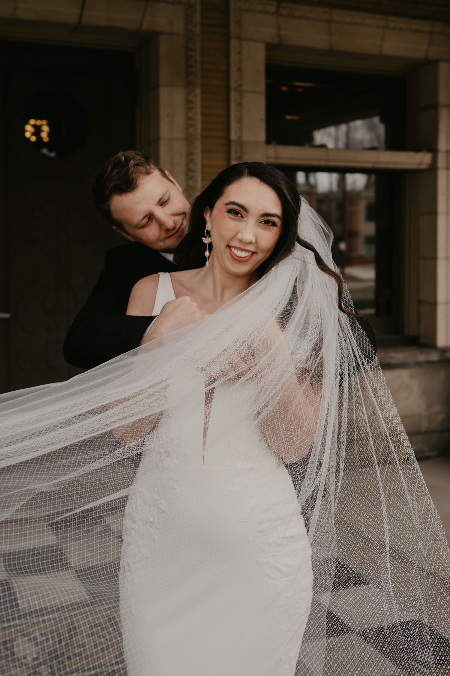 A radiant bride smiles as her groom adjusts her flowing veil outside a stone building.