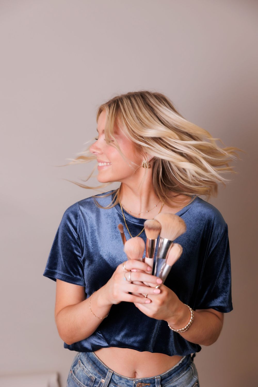 Woman with blonde hair flips her hair while holding makeup brushes and wearing blue velvet top.