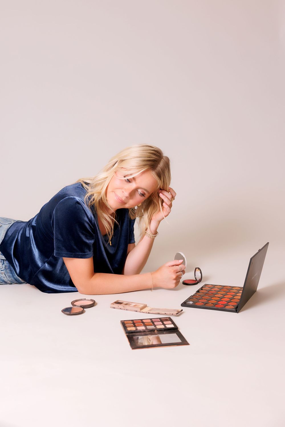 Person lying down with makeup palettes and laptop on light background.