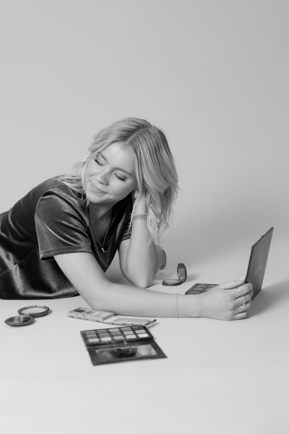 Woman in dark shirt lies on floor looking at laptop with makeup palette nearby.