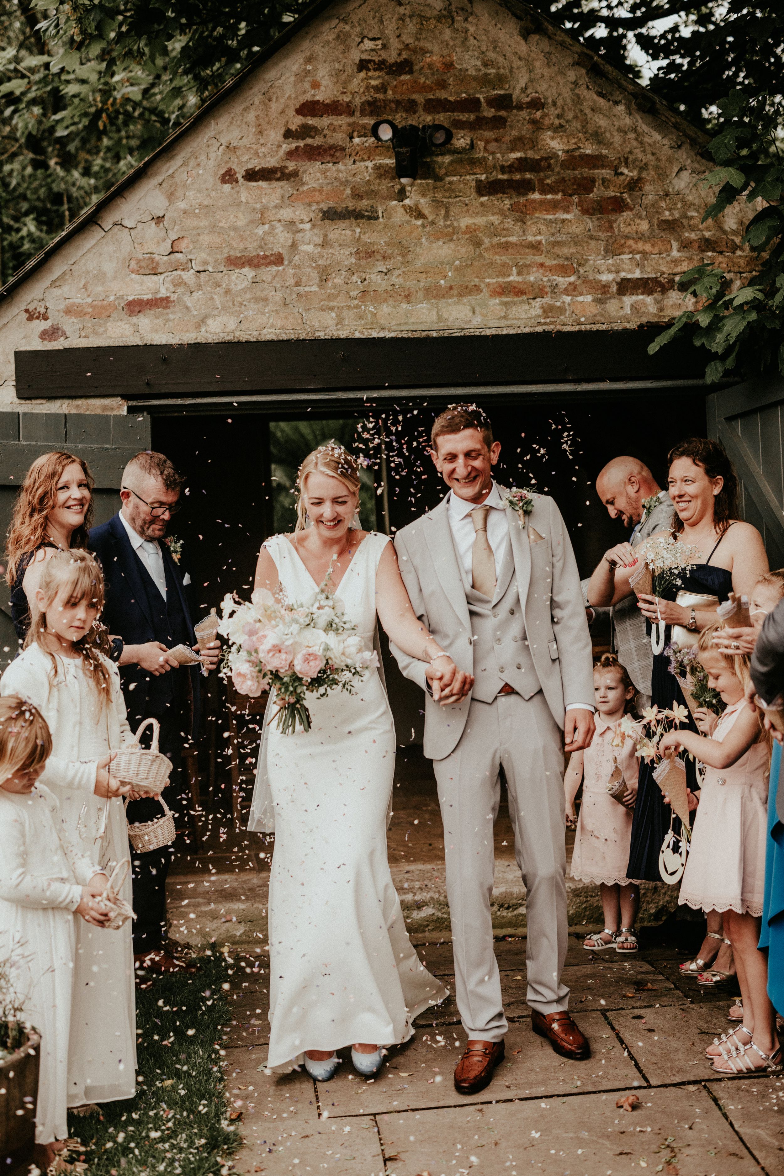 Bride and groom walk through confetti shower outside rustic brick venue, surrounded by smiling guests and flower girls.