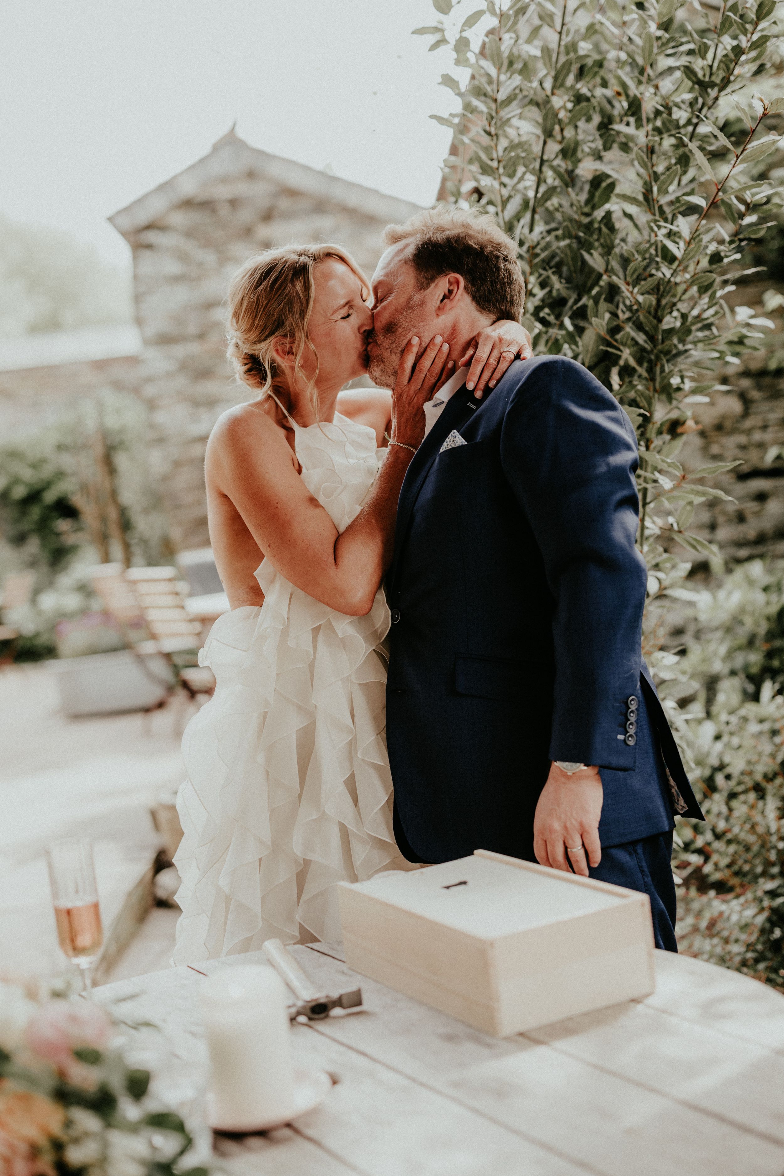 A bride and groom share a romantic kiss outdoors at their wedding reception, dressed in a white gown and navy suit.