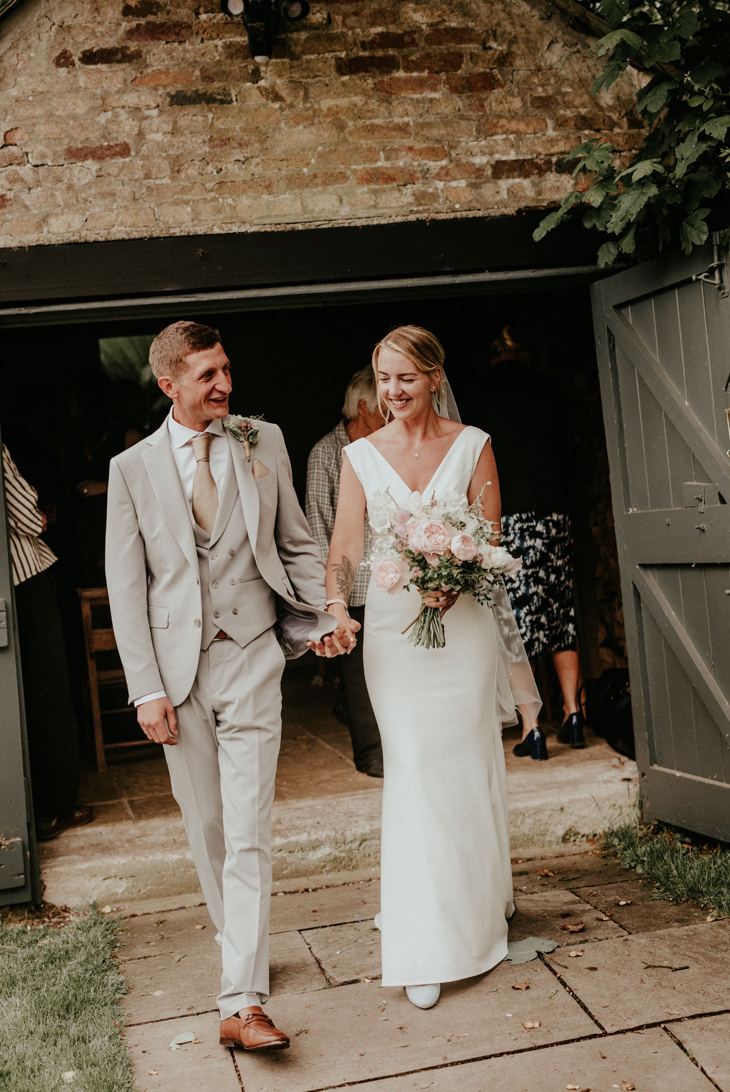 Smiling bride and groom holding hands walking out of rustic barn venue, bride in white gown holding pink bouquet.