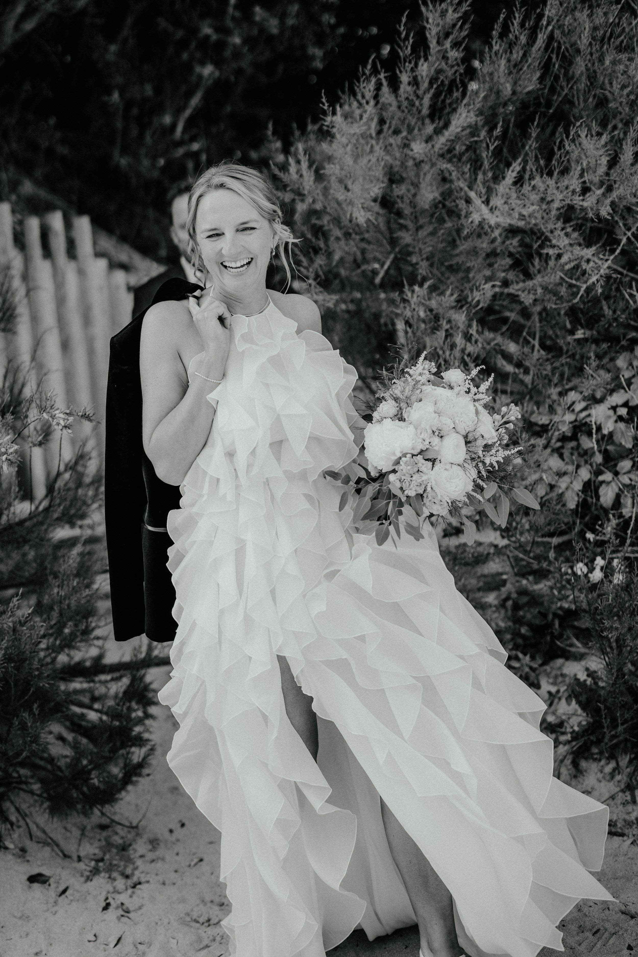 Black and white photo of a laughing bride in a ruffled gown holding a bouquet outdoors.